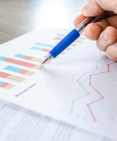 A close-up of a hand with a pen analyzing data on colorful bar and line charts on paper.